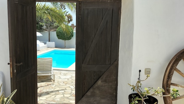 Una puerta que da a una zona de piscina con un patio de piedra, sillas reclinables blancas y un pequeño árbol.