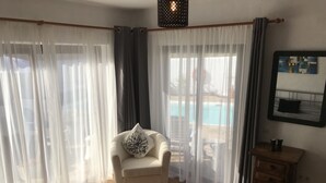 Un dormitorio con cama, una silla con cojín, un aparador de madera y vista a una piscina a través de cortinas translúcidas.