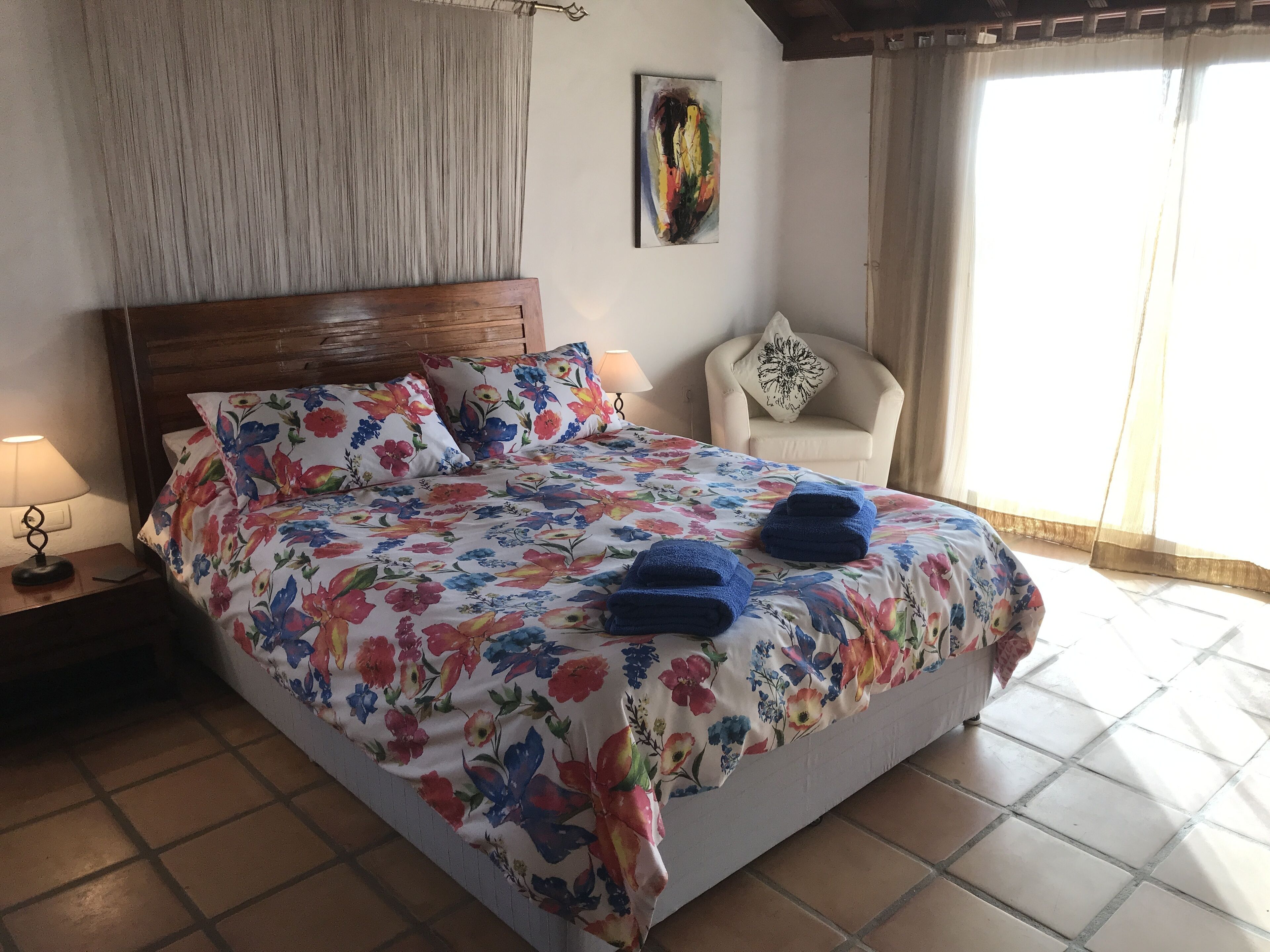 Een keurig opgemaakt bed met bloemenbeddengoed, een stoel met een decoratief kussen, en een schilderij aan de muur.