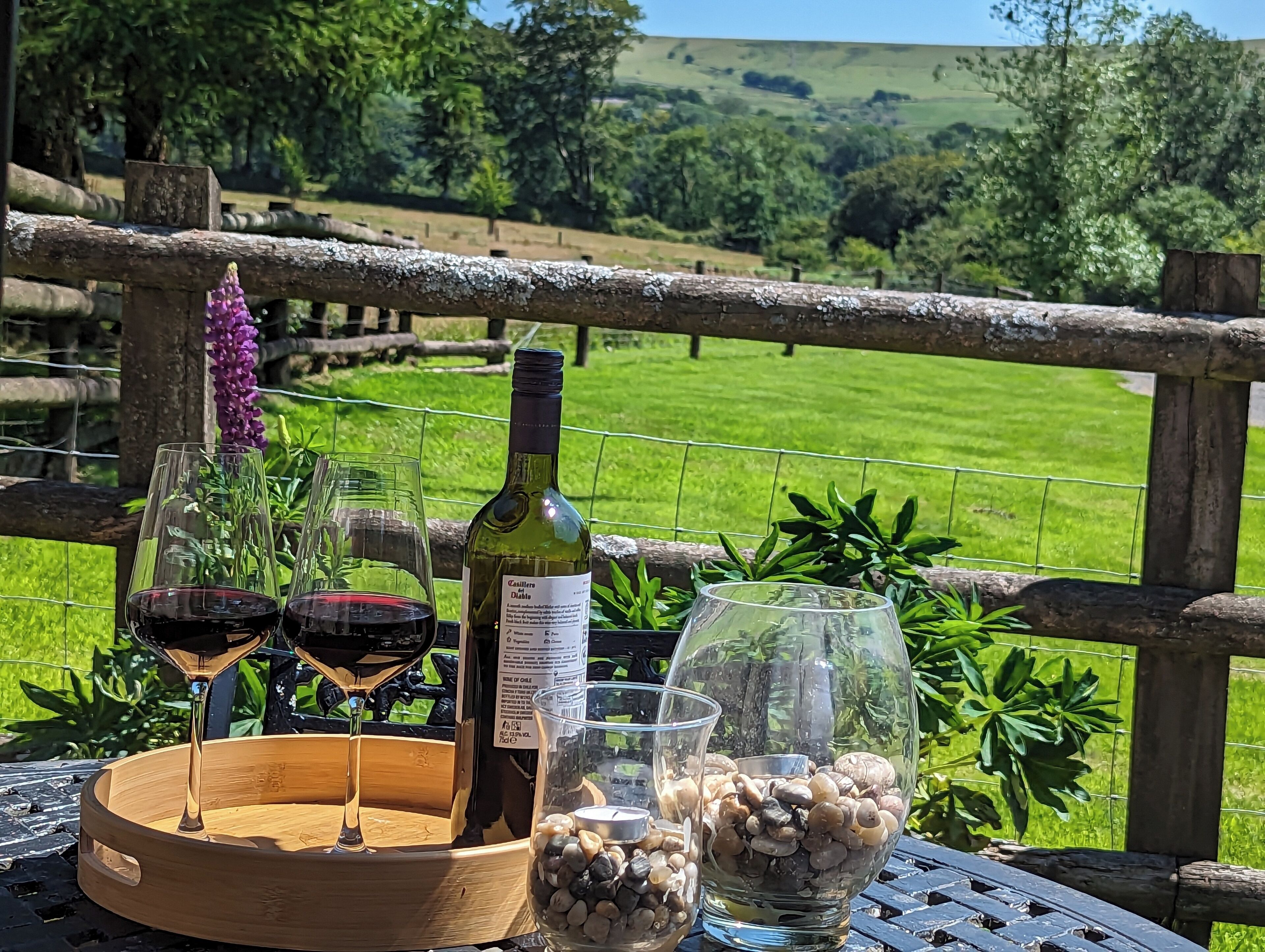 A table with a bottle of wine, two glasses of red wine, a glass container with pebbles, and a purple flower in the background.