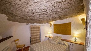 Une chambre rustique avec un plafond en pierre, un lit recouvert d’une couette à rayures, une chaise en bois, une table de chevet avec une lampe et une fenêtre avec un rideau.
