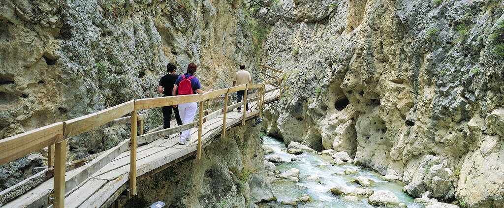Un pont en bois, sur lequel des personnes se promènent, mène à une falaise rocheuse au-dessus d’une rivière.