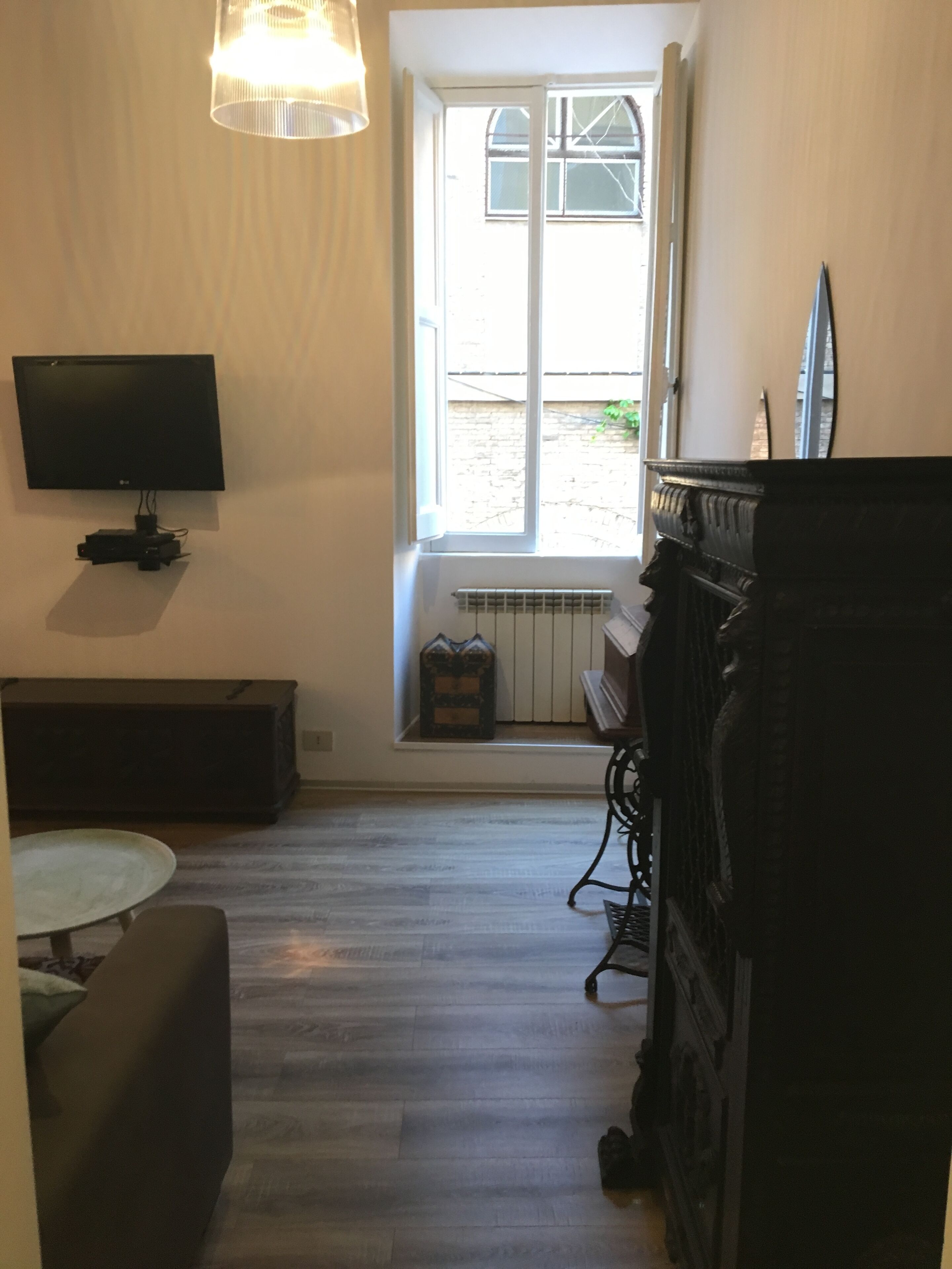 A room with a television mounted on the wall, a window with a view of the outside, a radiator below the window, a piano, and a table with a chair.