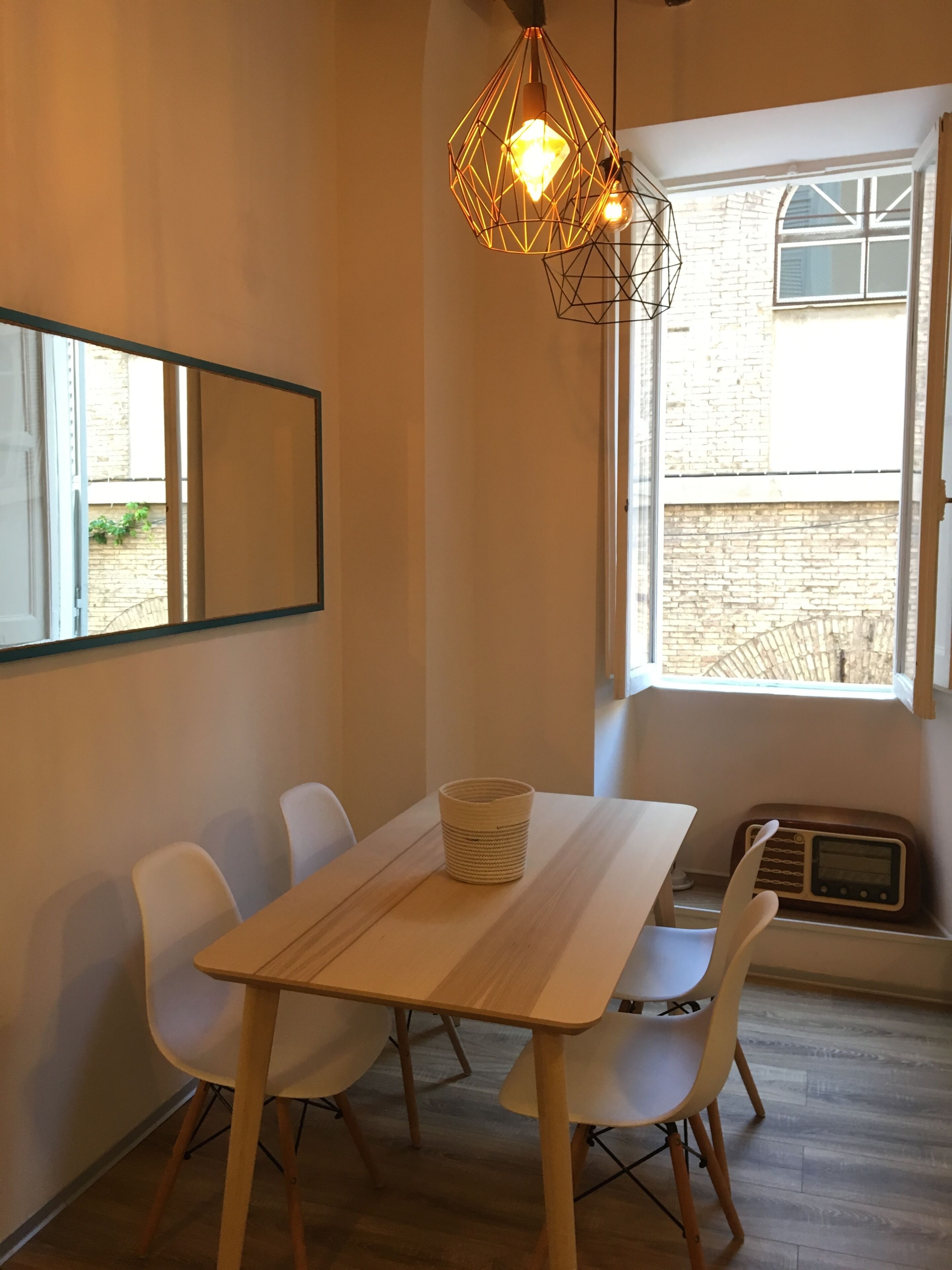 A dining area with a table and chairs, a hanging light fixture, a radio, and a window with a view of a brick building.