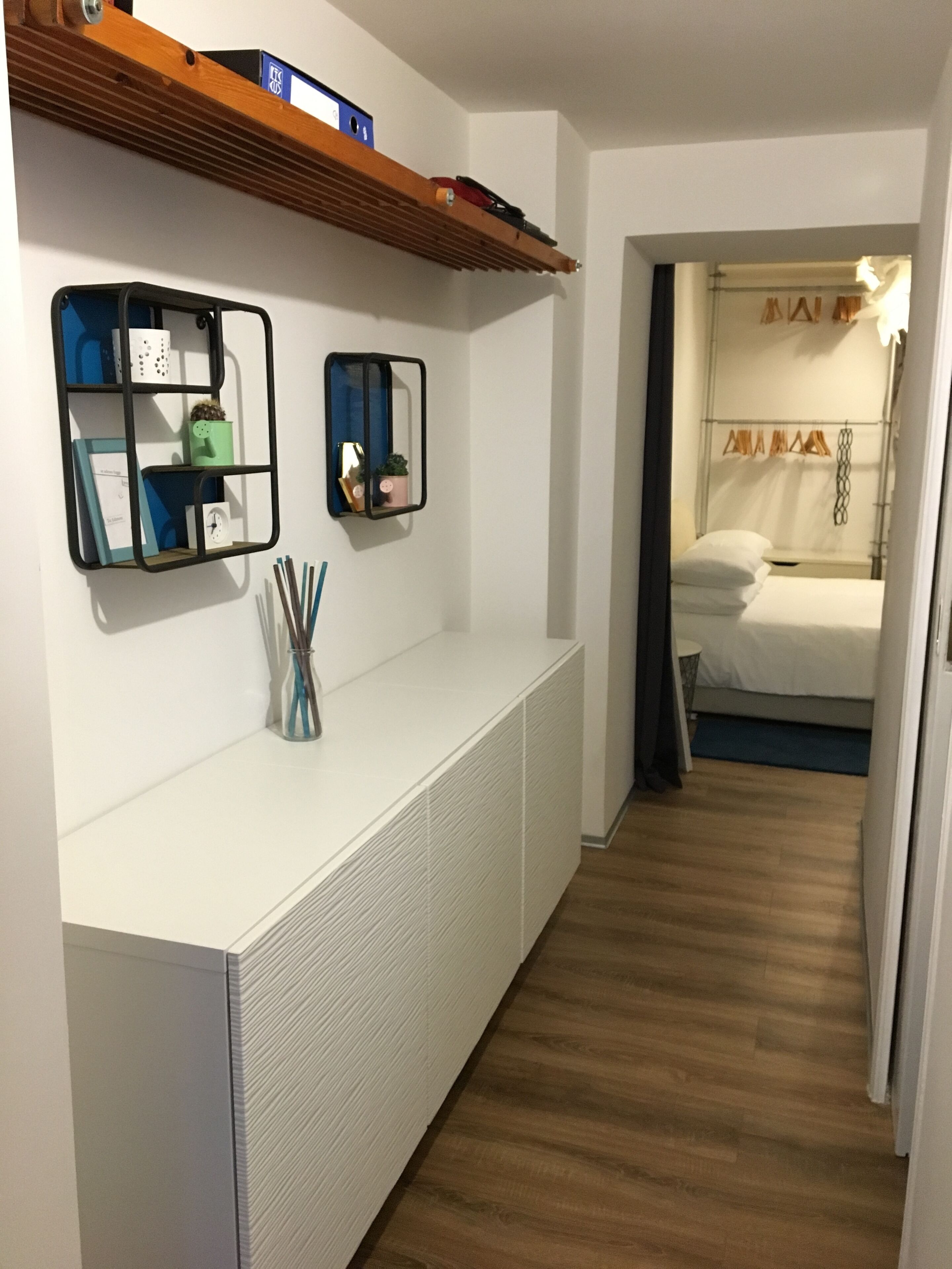 Habitación de hotel con una consola blanca, dos estantes fijos decorados y un suelo de madera, además de una cama con ropa de cama blanca.