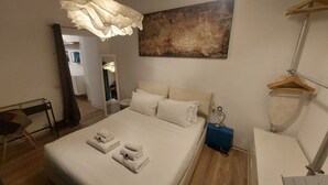 Un lit bien fait, avec du linge de lit et des serviettes blanches, une table de chevet avec une lampe, et un grand tableau abstrait accroché au mur.
