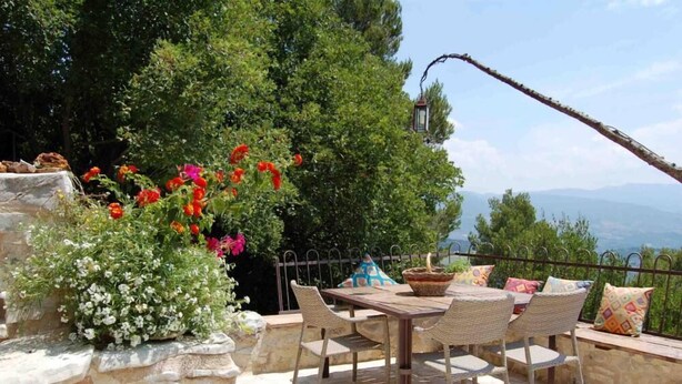 Eine Terrassengruppe im Freien mit einem Steintisch und Korbstühlen, umgeben von üppiger Bepflanzung und Blick auf entfernte Berge.