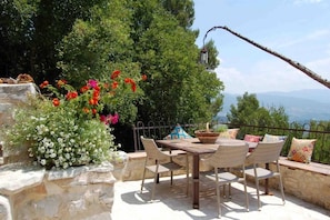 An outdoor patio with a stone table and wicker chairs, surrounded by lush greenery and a view of distant mountains.