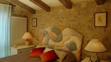 A bedroom with a stone wall, a bed with a patterned headboard, two bedside lamps, and a window with curtains.