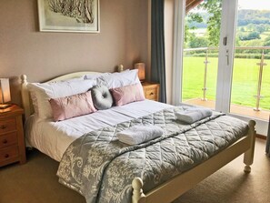 A bedroom with a bed, a nightstand, a lamp, a window with a view of greenery, and a painting on the wall.