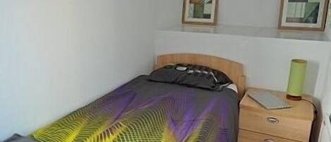 Un lit simple avec une couvre-lit à motifs, une table de chevet avec une lampe et deux tableaux encadrés accrochés au mur.