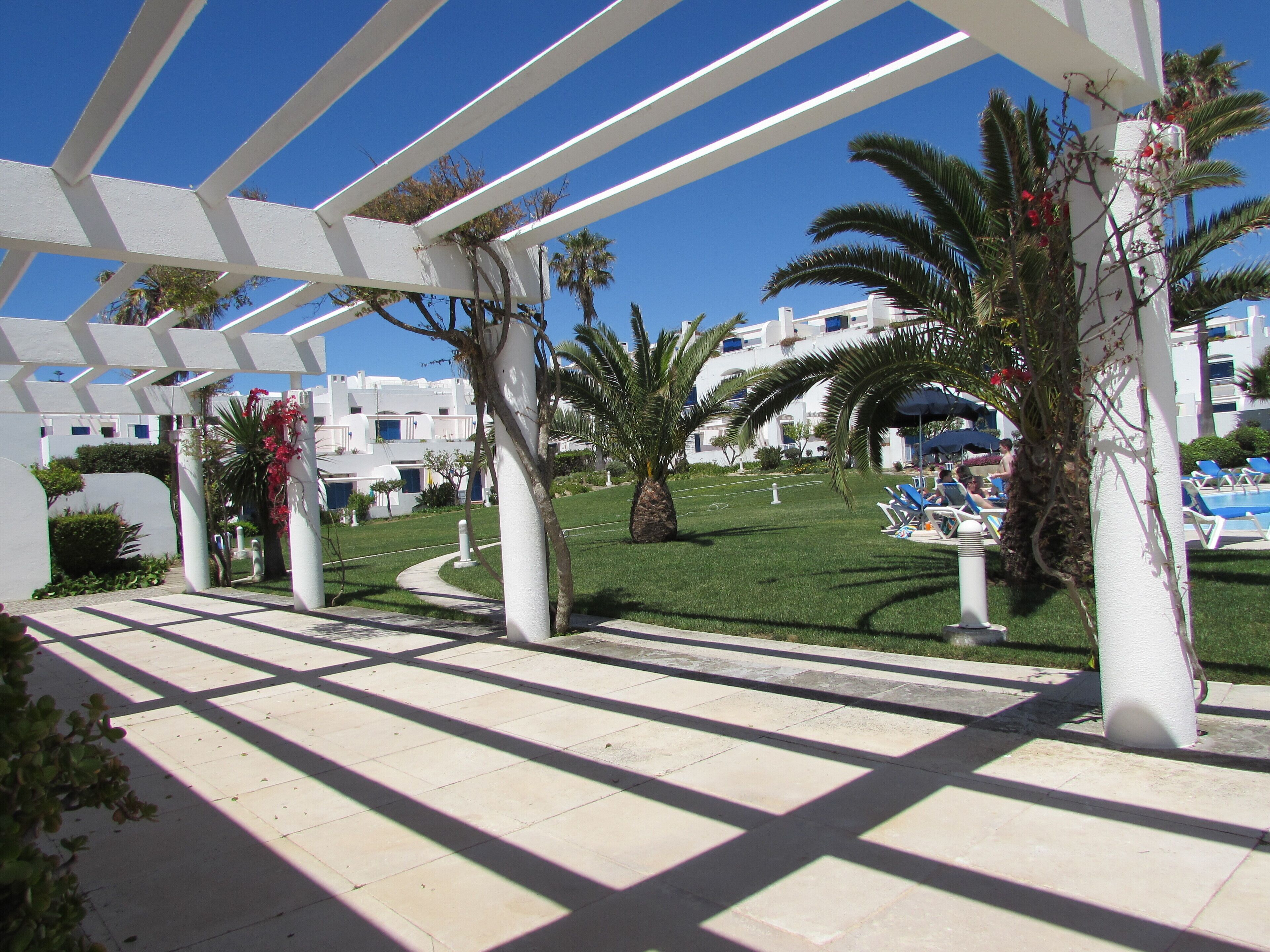 A covered outdoor area with a tiled floor, palm trees, and a view of a building with multiple balconies.