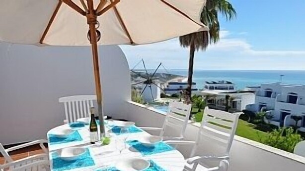 Et rundt bord med hvide stole og en beige parasol, placeret på en balkon med udsigt over kysten med hvide bygninger og en vindmølle.