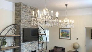 Ein Wohnzimmer mit Kamin aus Stein, einer großen Chandelierleuchtre, einem schwarzen Ledersofa und einem an der Wand befestigten Fernseher.