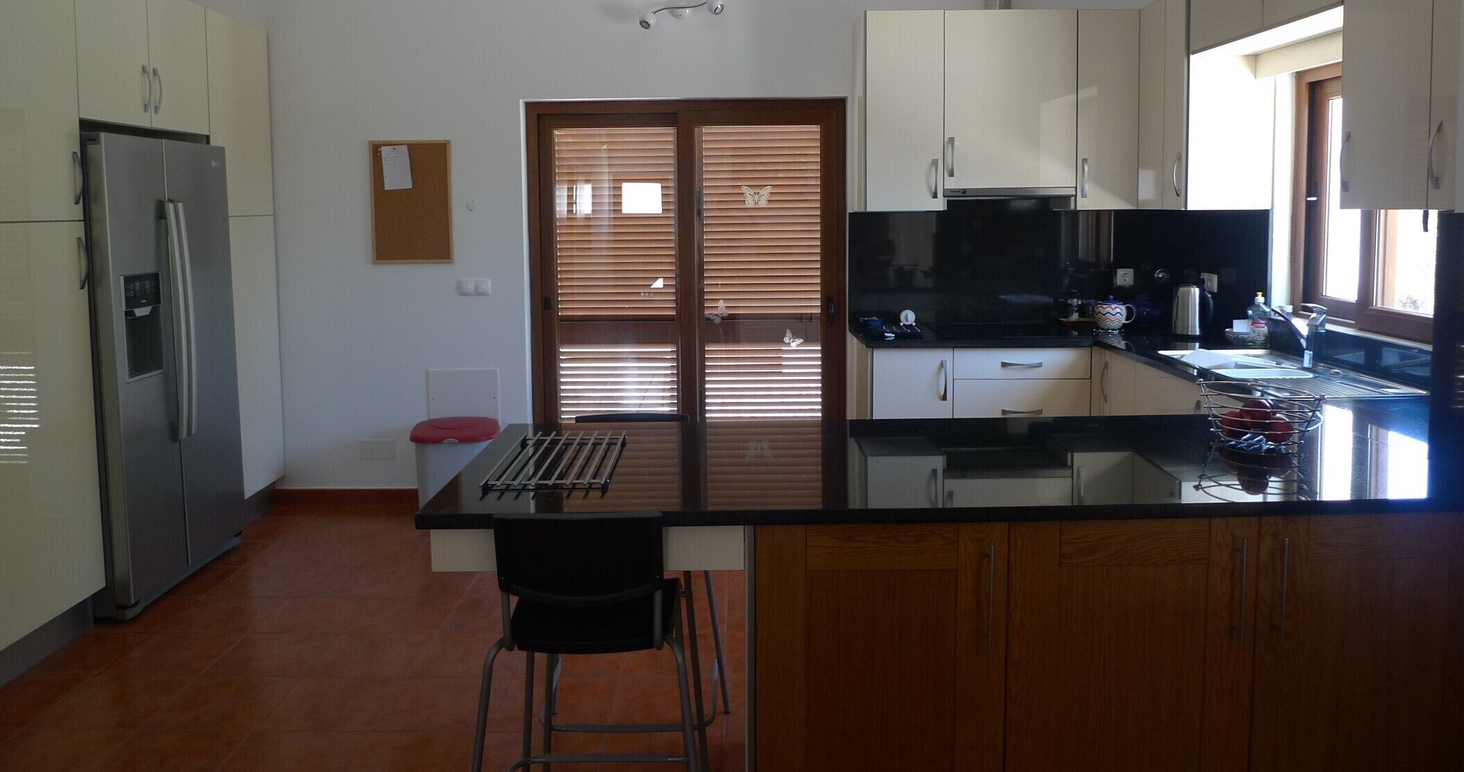 A modern kitchen with a stainless steel refrigerator, wooden cabinets, a black countertop, and a window with blinds.