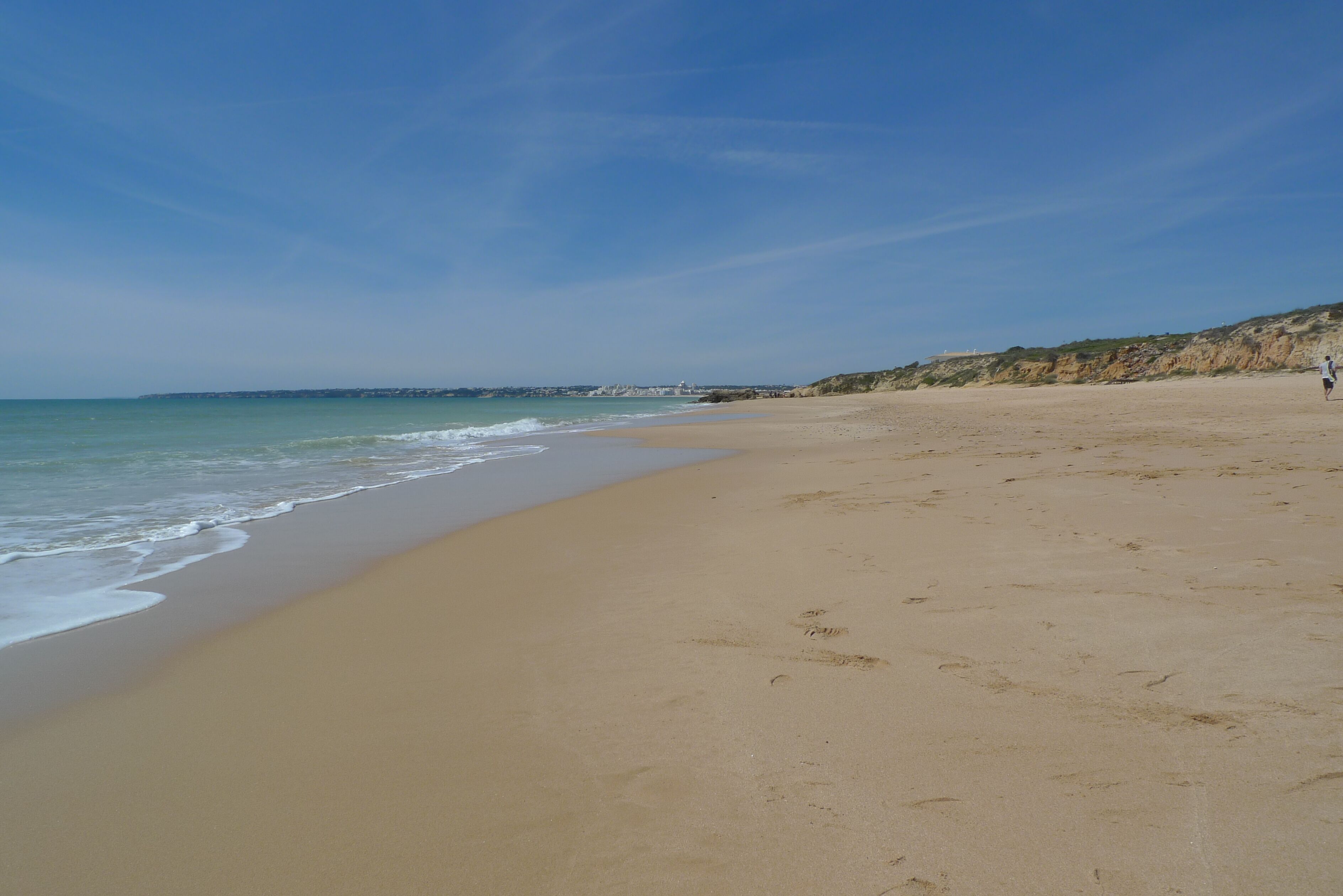 A wide sandy beach with gentle waves, a clear sky, and a distant coastline.