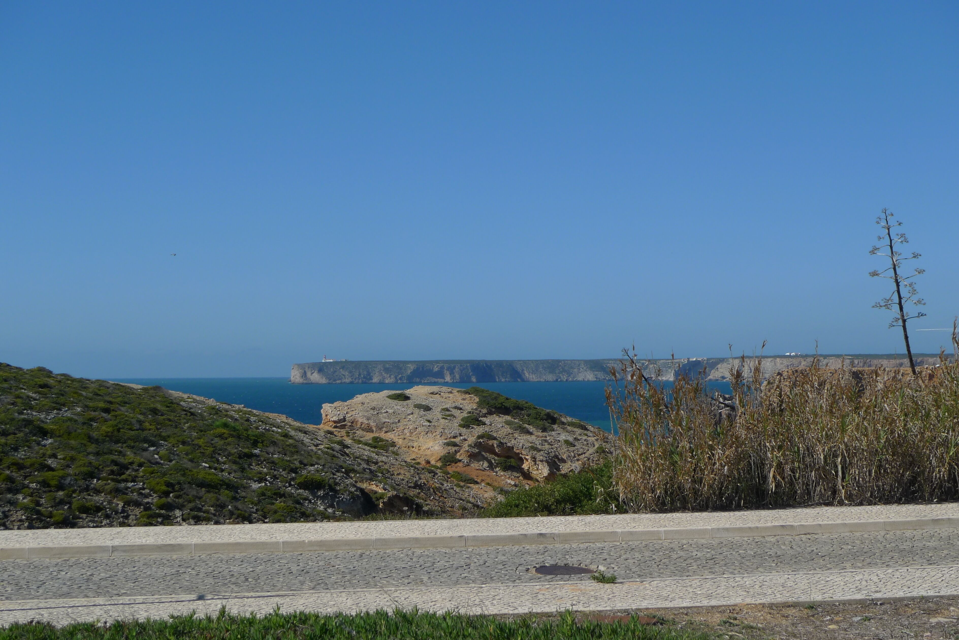 Une route côtière traversant un paysage rocheux, sous un ciel bleu dégagé, avec une vue lointaine sur des falaises et la mer.