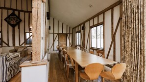 Una larga mesa de madera con sillas en una habitación rústica con vigas a la vista y ventanas amplias.