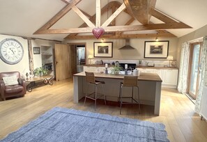 A spacious kitchen with a central island, wooden beams, a clock, and a dining area with a chair and a table. - Houses and Grounds of Partridge Lodge (Woodbridge)