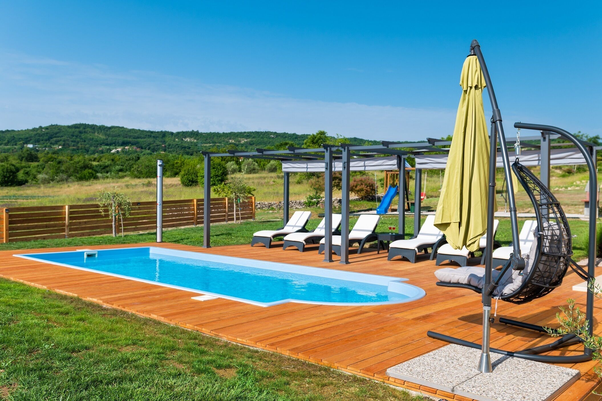 Une piscine entourée de chaises longues et d’une chaise suspendue, le tout bordé d’une terrasse en bois et surmonté d’une pergola avec un parasol jaune.