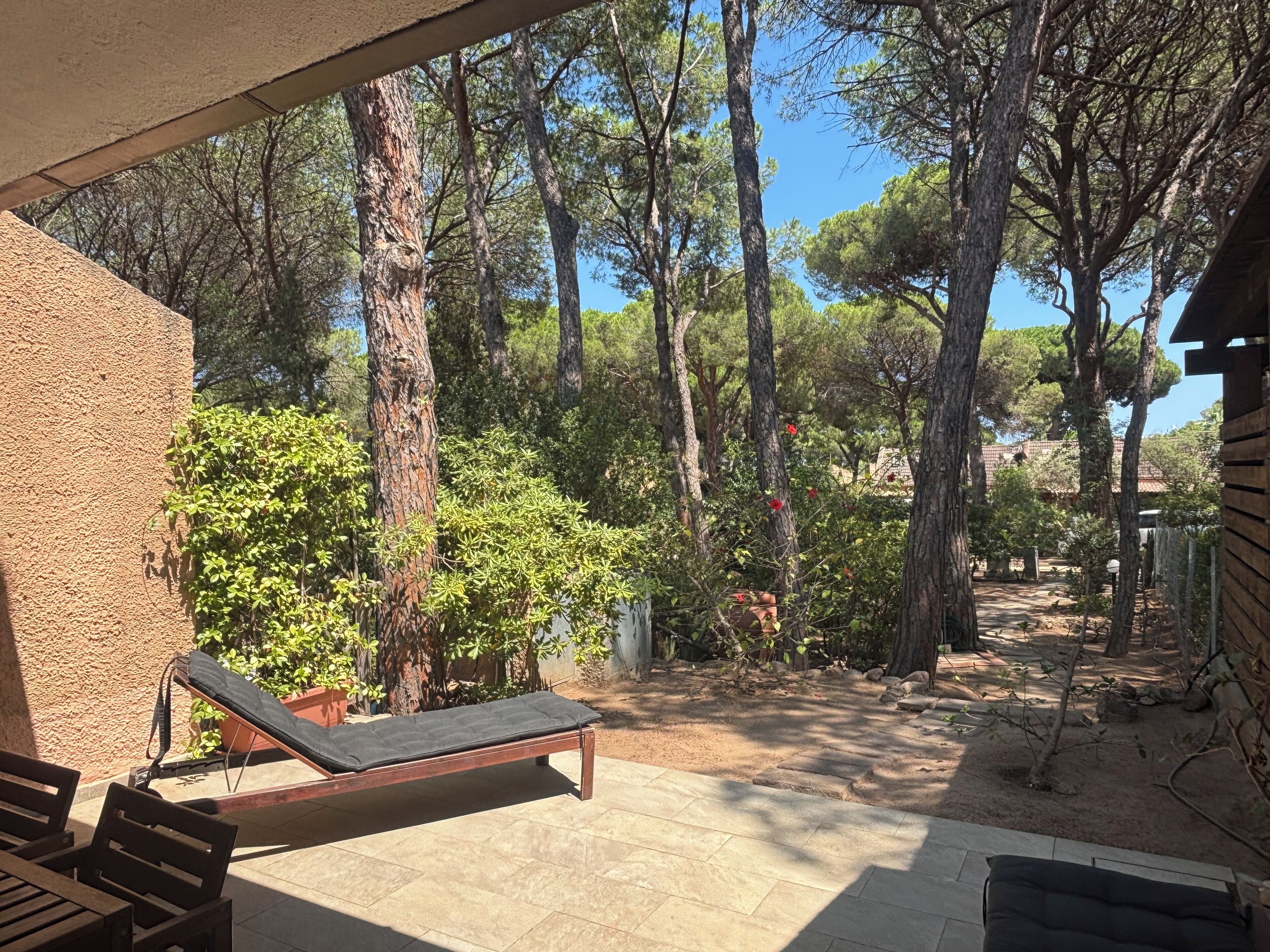 A covered outdoor area with a lounge chair and a bench, surrounded by trees and a clear sky.