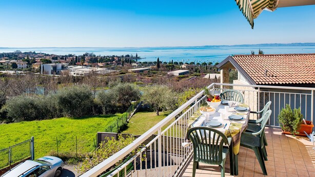 A balcony with a table set for dining, overlooking a green landscape and a distant body of water. - Relax overlooking Lake Garda (Bardolino)