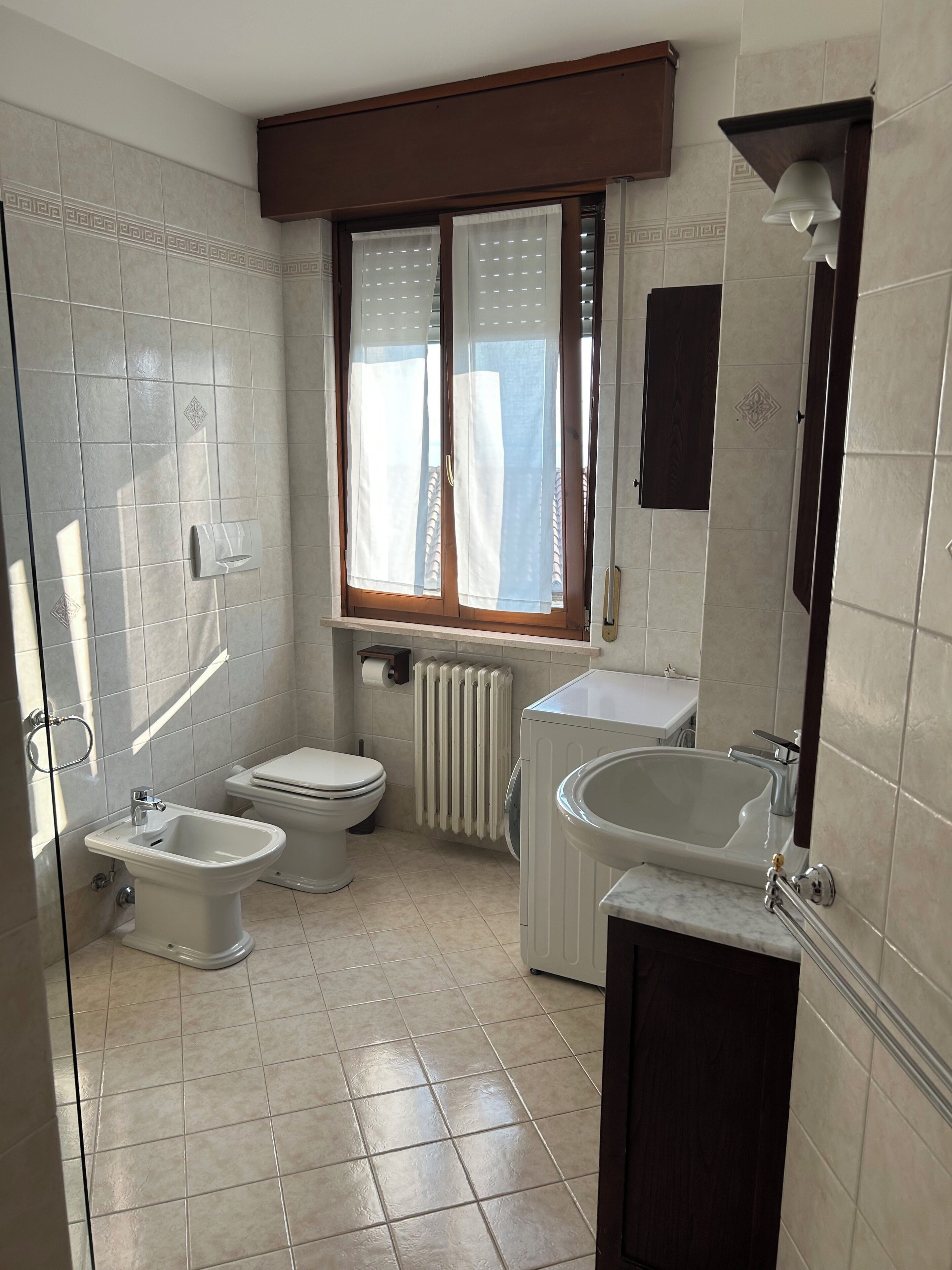 Une salle de bain comprenant des toilettes, un bidet et un lavabo, une fenêtre avec des rideaux et un radiateur.