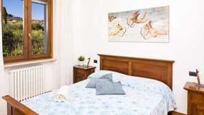 A bedroom with a wooden bed, a view of the outdoors through a window, a painting on the wall, and a bedside table with a lamp. - Relax overlooking Lake Garda (Bardolino)