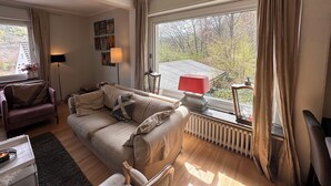 Ett mysigt vardagsrum med en beige soffa, ett litet bord, en golvlampa och ett element nära ett stort fönster med gardiner.