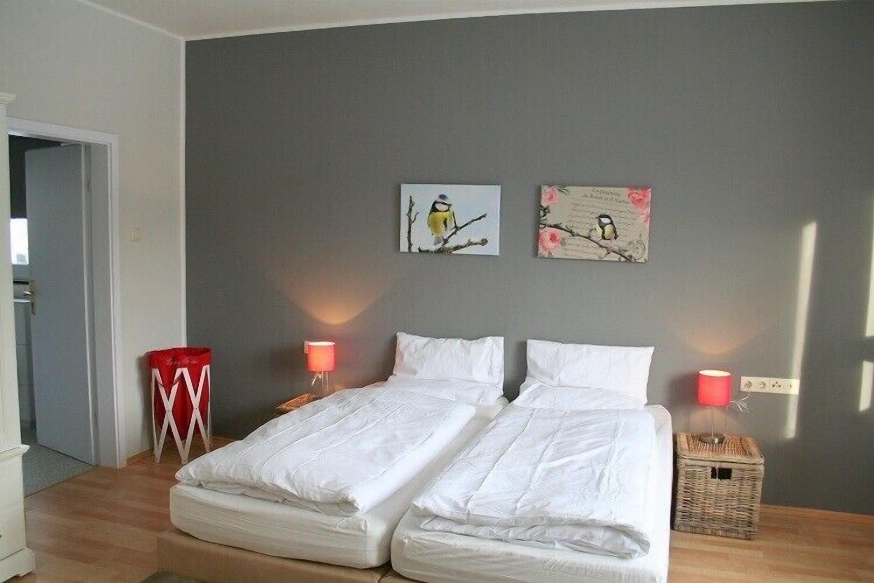 A room with two single beds, a red chair, a small table, and two wall-mounted lamps.