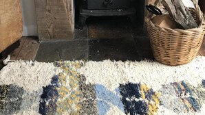 A wood-burning stove with a "Welcome" sign above it, a woven basket with logs, and a patterned rug on the floor.
