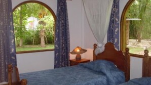 Ein Schlafzimmer mit zwei Betten, darunter ein Himmelbett, einem Nachttisch mit Lampe und Blick auf Grün durch gewölbte Fenster.