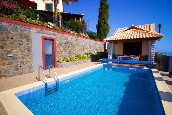 A swimming pool with a stone wall and a door, surrounded by a terrace with plants and a view of the sea.