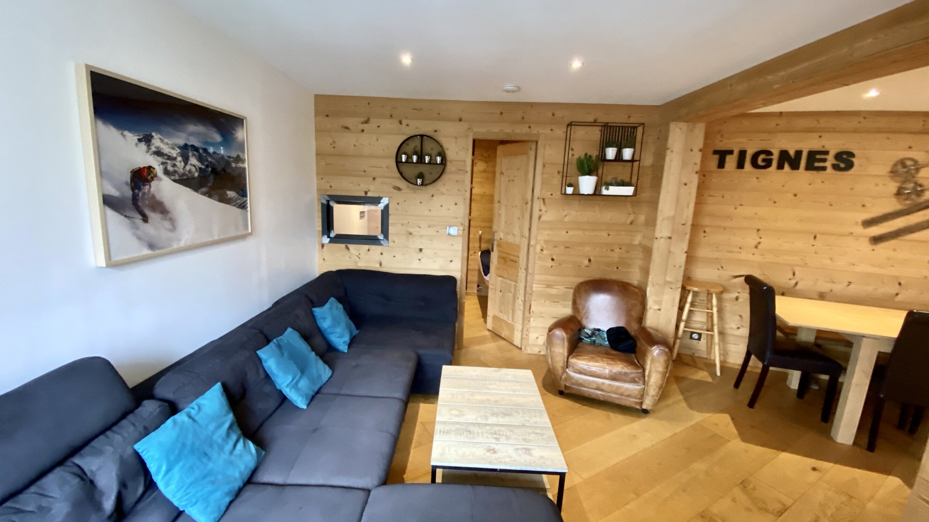 A modern living room with a sectional sofa, a coffee table, a leather armchair, and a dining area with a wooden wall and a sign reading 'TIGNES'.