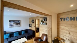 Een moderne woonkamer met een donkere bank, een salontafel en een leren fauteuil. Er is een houten wand met een plank waarop planten staan en een bord met de tekst 'TIGN'.