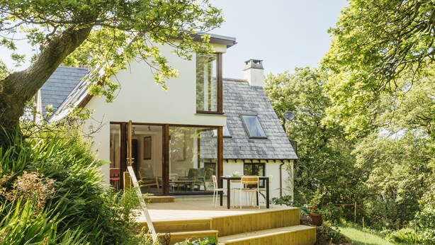 A modern house with a sloped roof, a wooden deck, and a glass door leading to a patio. - Contemporary cottage retreat in secluded valley near sea. Sleeps 8 (Bude)