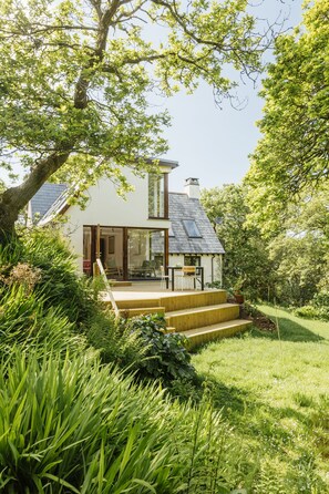 A modern house with a sloped roof, a wooden deck, and a glass door leading to a patio. - Contemporary cottage retreat in secluded valley near sea. Sleeps 8 (Bude)