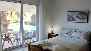 Une chambre d’hôtel avec un lit, une table de chevet avec une lampe, un tableau au mur et une vue sur une piscine extérieure et des chaises de patio, visibles à travers la porte-fenêtre coulissante.