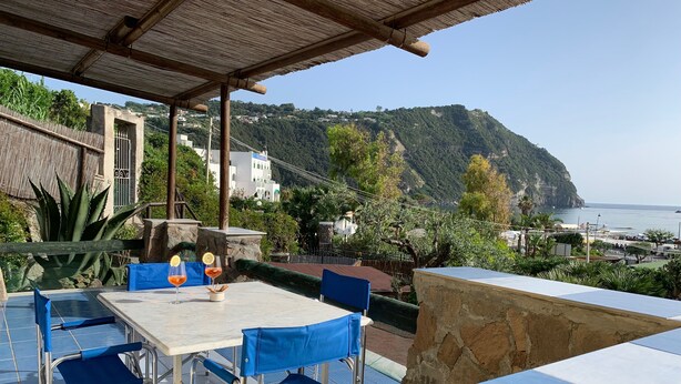 Une terrasse avec une table et des chaises, offrant une vue sur un paysage côtier avec des maisons et une montagne.