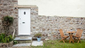A stone building with a white door, a small garden with a potted plant, and two wooden chairs around a table with a bottle. - Bay Cottage, a section of Ebbingford Manor, 850 yr old manor house by beach&town (Bude)