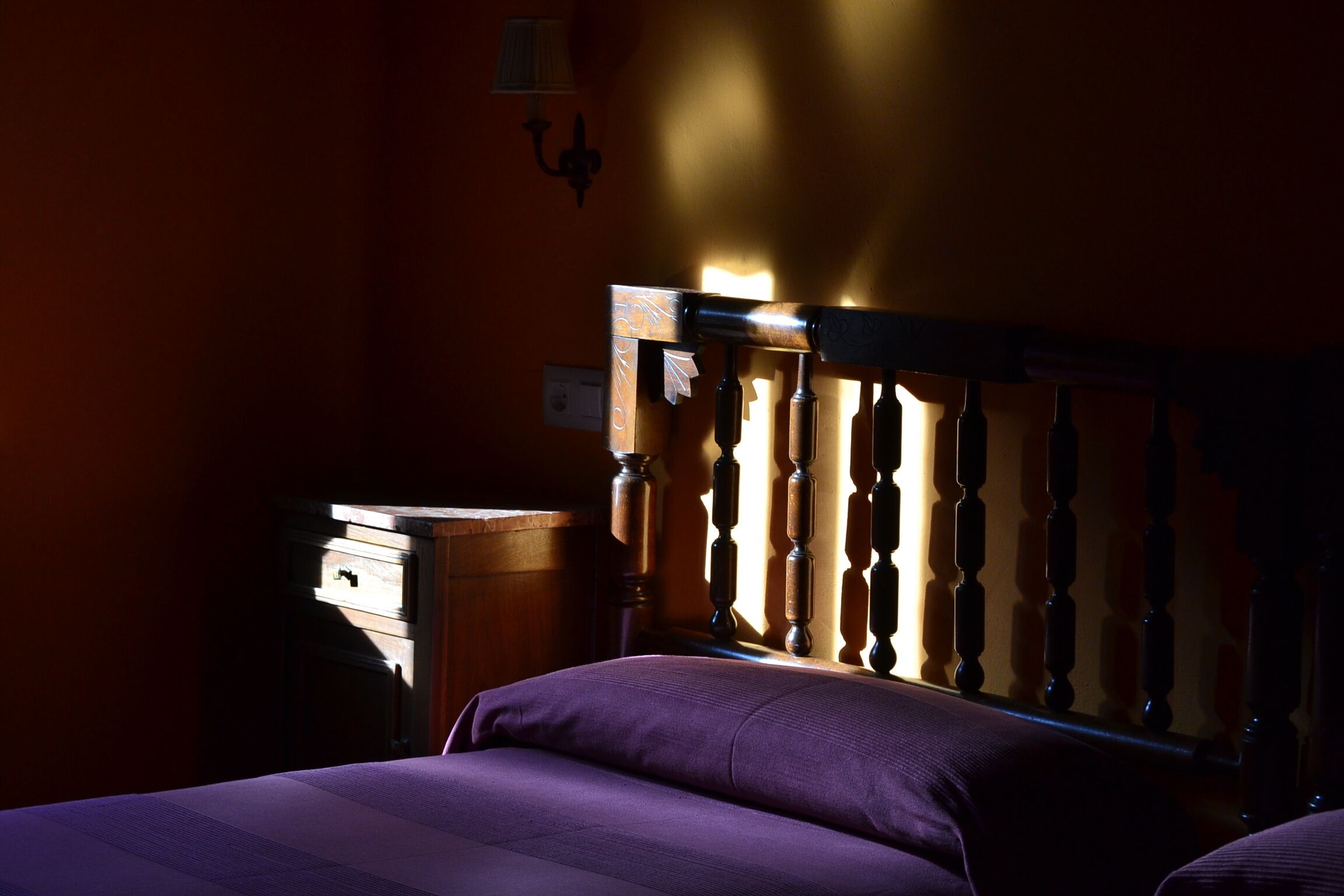 A room with a bed, a wooden headboard, a nightstand, and a wall-mounted lamp.