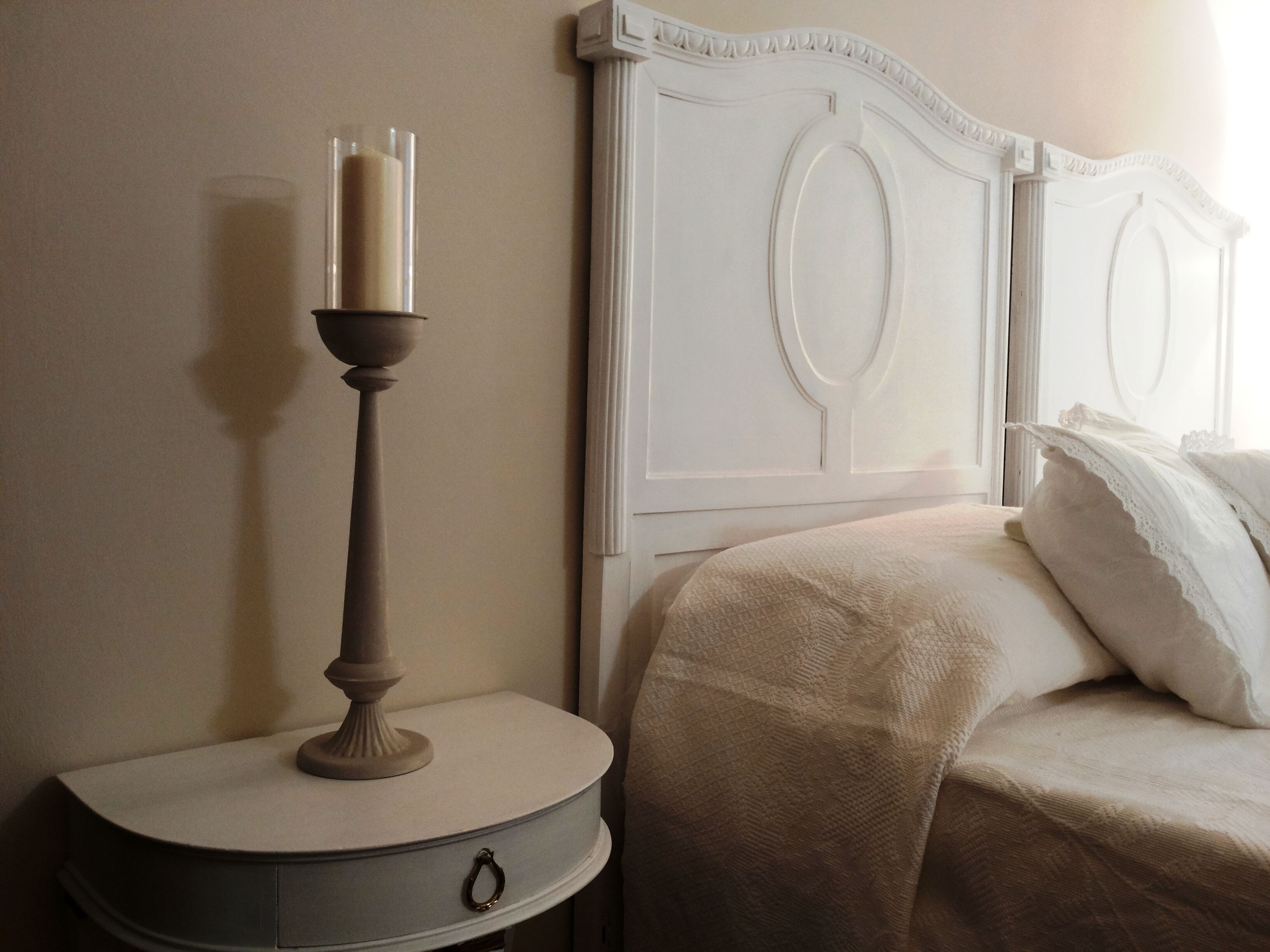 Un lit bien fait, avec une tête de lit blanche, une table de chevet sur laquelle se trouve un chandelier et un petit tiroir.