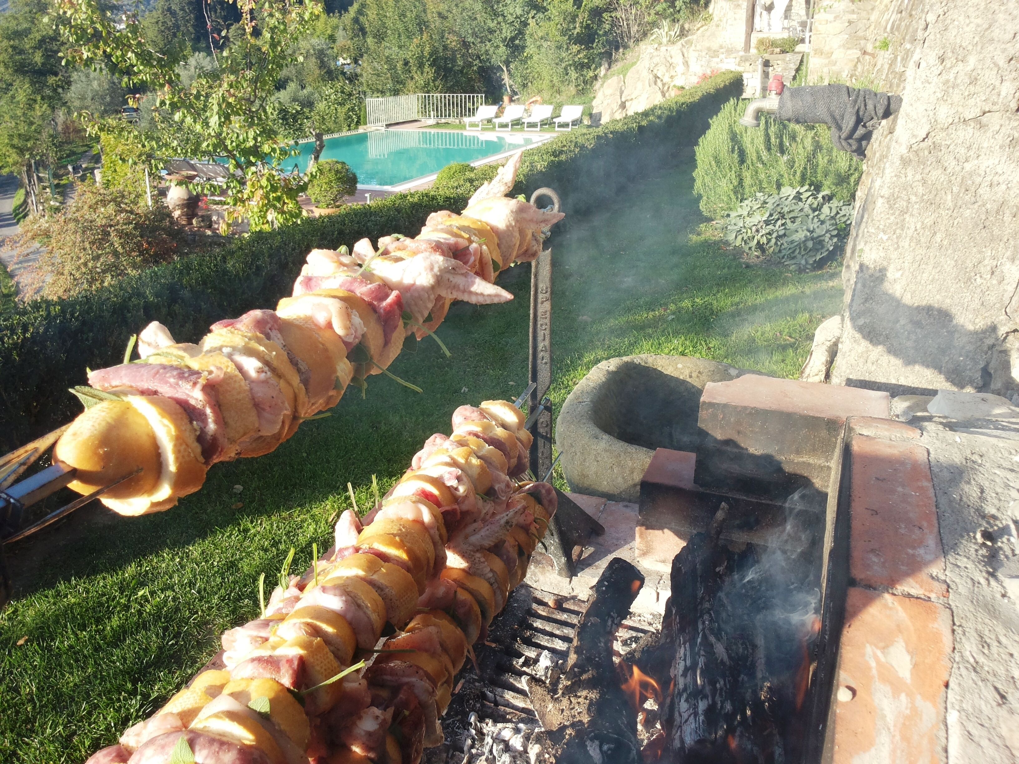 A spit-roasted meat skewer over an open fire, with a pool and greenery in the background.