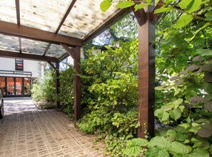 A covered outdoor area with a car parked under a metal roof, surrounded by greenery and a brick pathway leading to a building entrance.