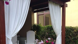 A covered patio area with white curtains, a white chair, and a large planter with pink flowers. - Villa with garden just 200 meters from the beautiful beach of Costa Rei (Costa Rei)