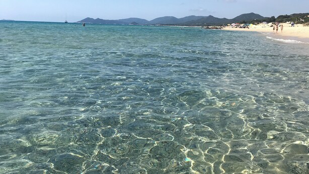 A beach with clear shallow waters, distant mountains, and a few people walking along the shore. - Villa with garden just 200 meters from the beautiful beach of Costa Rei (Costa Rei)