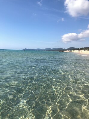 Ein Strand mit klarem, flachem Wasser, Bergen im Hintergrund und einigen Personen, die am Ufer entlanggehen.
