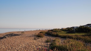 A pebbled beach with boats in the water, a golf course, and houses in the background. - Beach front location, Pet friendly, Full of charachter, Private garden, Sea view (Selsey)