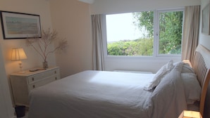 A bedroom with a large bed, a nightstand with a lamp, a framed picture on the wall, and a window with curtains. - Beach front location, Pet friendly, Full of charachter, Private garden, Sea view (Selsey)
