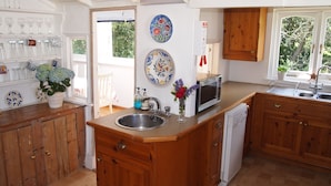 A kitchen with wooden cabinets, a central island with a sink, a microwave, and a dish rack. There are decorative plates on the wall and a potted plant on the counter. - Beach front location, Pet friendly, Full of charachter, Private garden, Sea view (Selsey)